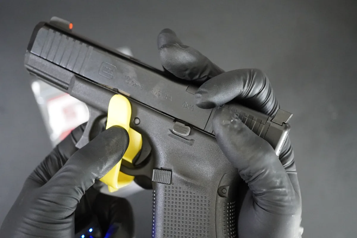Gloved hands pulling down both takedown levers on a Glock 19 Gen 5 while easing the slide forward, with a yellow chamber safety flag in place